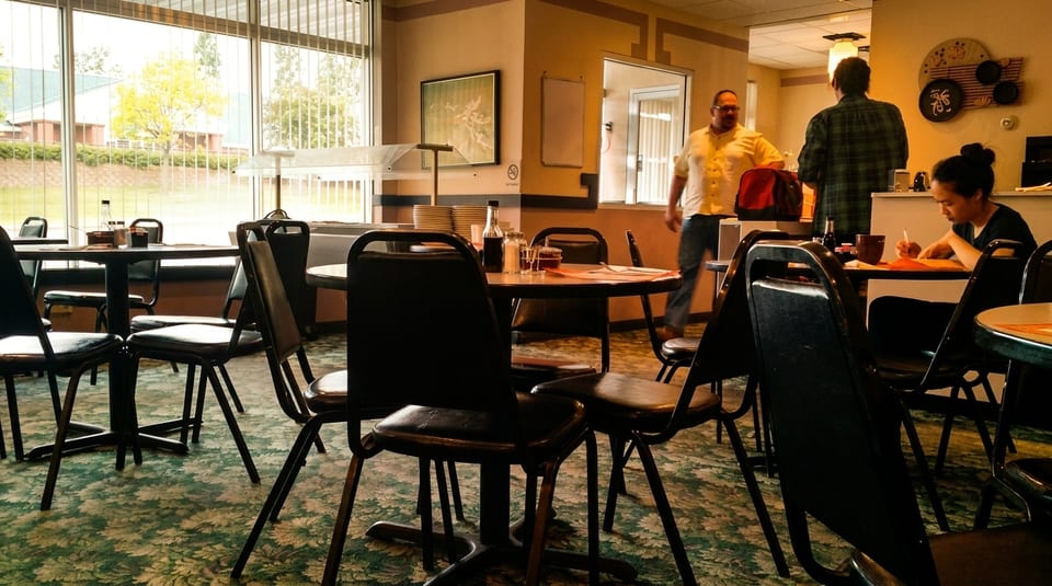 Restaurant interior with tables, chairs, a large window, and people walking and sitting.