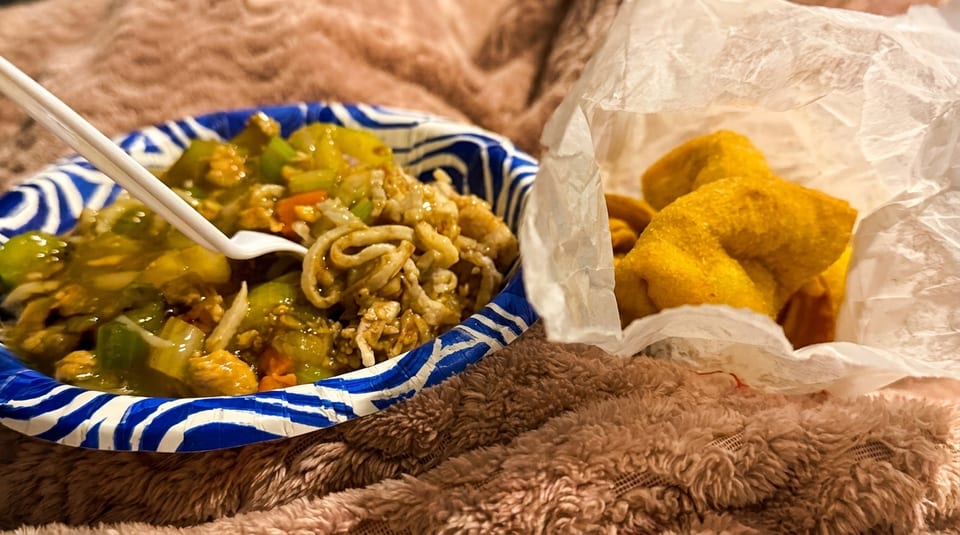 A paper plate of stir-fry with a fork and a bag of fried wontons on a fuzzy blanket.